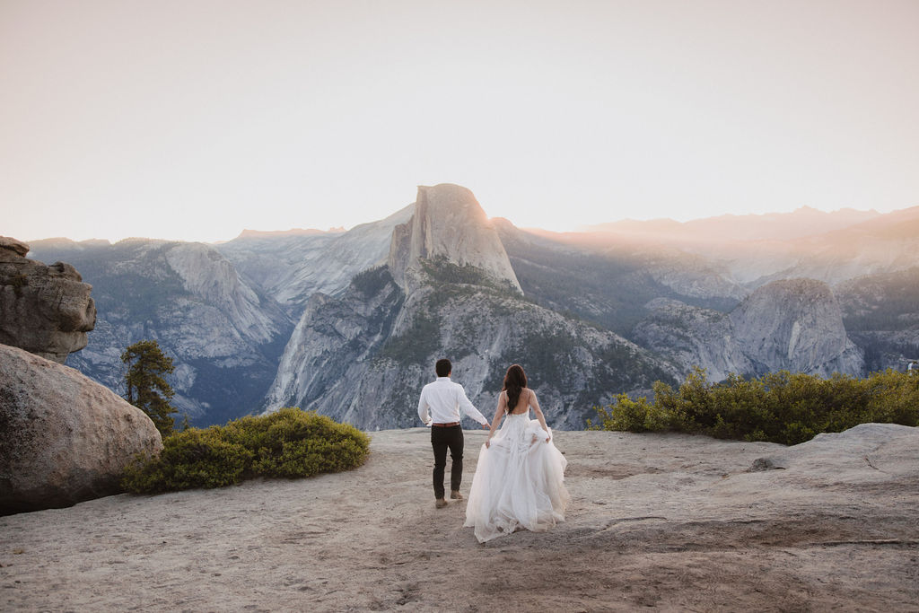 A couple stands on a rocky ledge during sunset; the man, wearing a white shirt and dark pants, holds the hand of the woman, who is in a flowing white dress. A mountain peak is visible in the background for their yosemite elopement photos