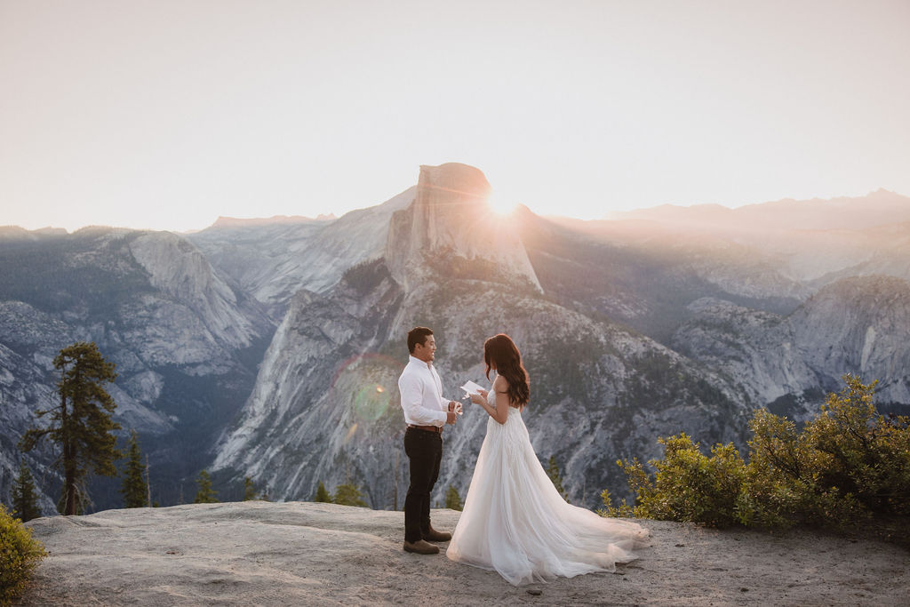 A couple stands on a rocky ledge during sunset; the man, wearing a white shirt and dark pants, holds the hand of the woman, who is in a flowing white dress. A mountain peak is visible in the background for their yosemite elopement photos