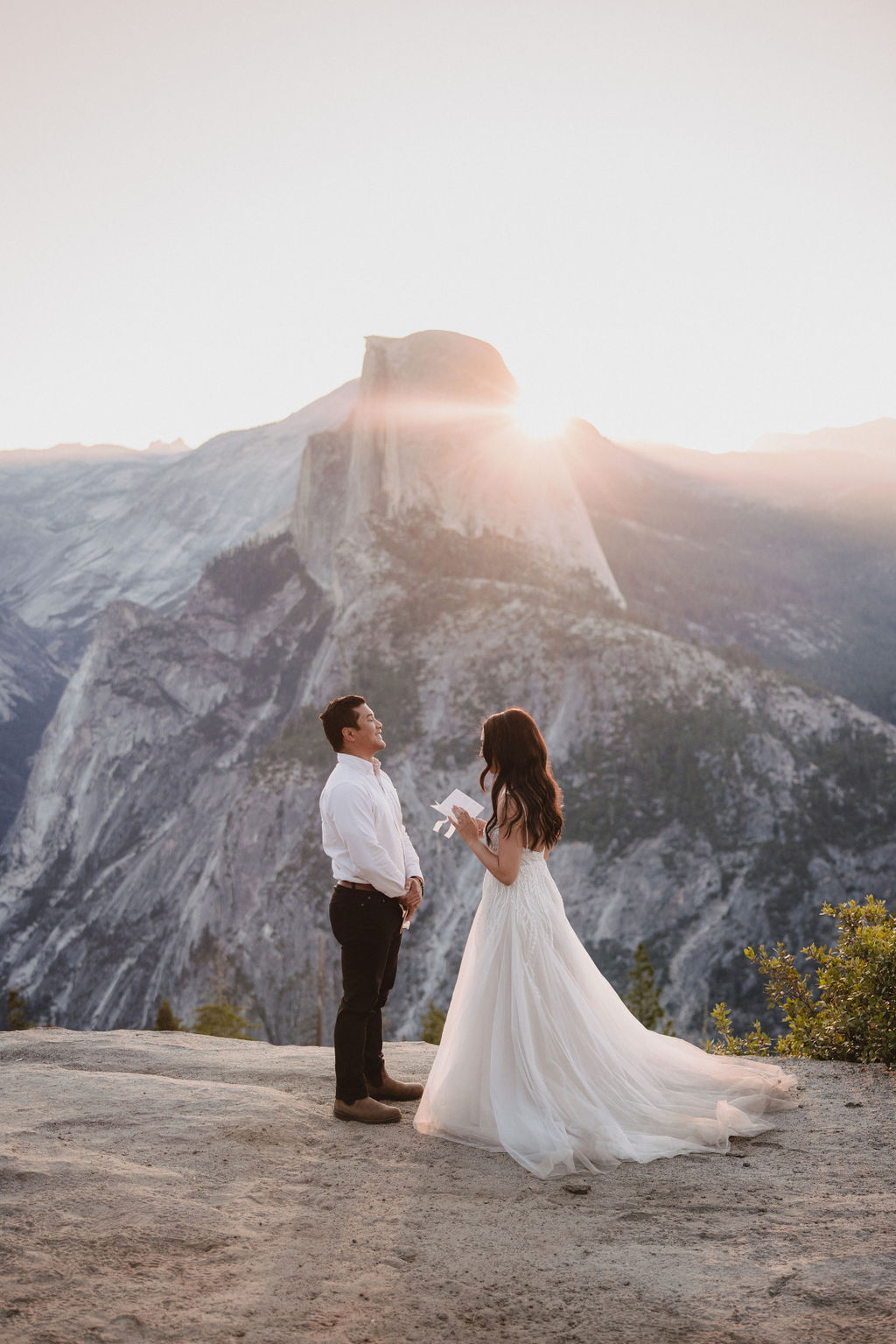 A couple stands on a rocky ledge during sunset; the man, wearing a white shirt and dark pants, holds the hand of the woman, who is in a flowing white dress. A mountain peak is visible in the background for their yosemite elopement photos