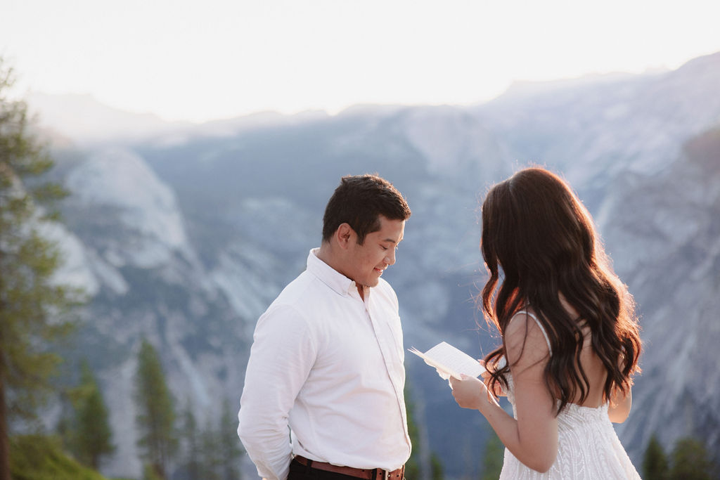 A couple stands on a rocky ledge during sunset; the man, wearing a white shirt and dark pants, holds the hand of the woman, who is in a flowing white dress. A mountain peak is visible in the background for their yosemite elopement photos