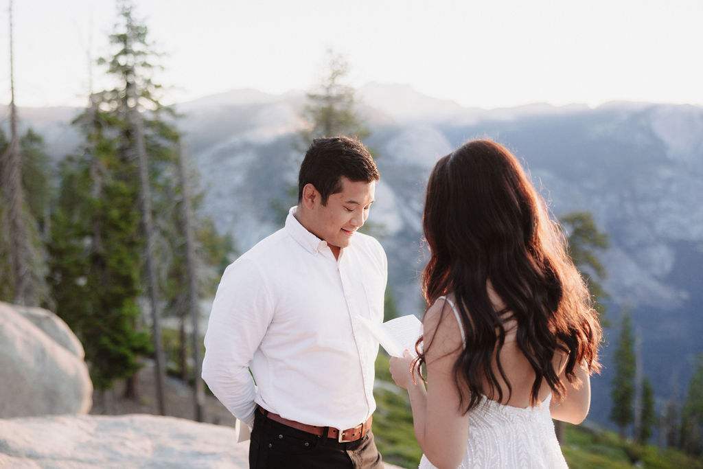 A couple stands on a rocky ledge during sunset; the man, wearing a white shirt and dark pants, holds the hand of the woman, who is in a flowing white dress. A mountain peak is visible in the background for their yosemite elopement photos