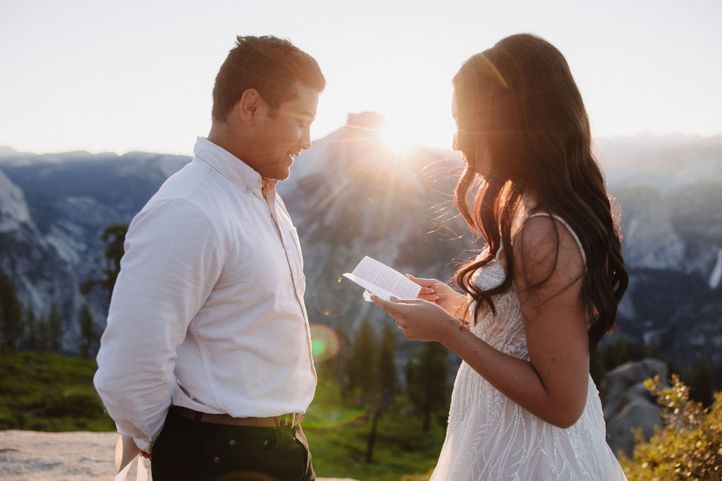 A couple stands on a rocky ledge during sunset; the man, wearing a white shirt and dark pants, holds the hand of the woman, who is in a flowing white dress. A mountain peak is visible in the background for their yosemite elopement photos