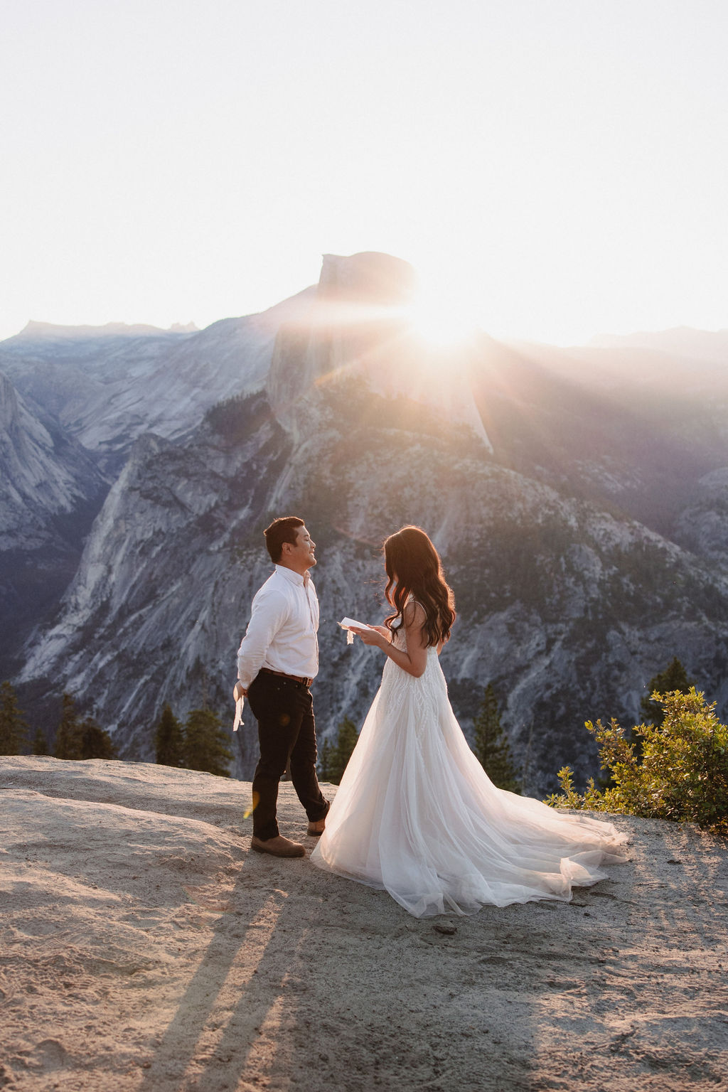 A couple stands on a rocky ledge during sunset; the man, wearing a white shirt and dark pants, holds the hand of the woman, who is in a flowing white dress. A mountain peak is visible in the background for their yosemite elopement photos