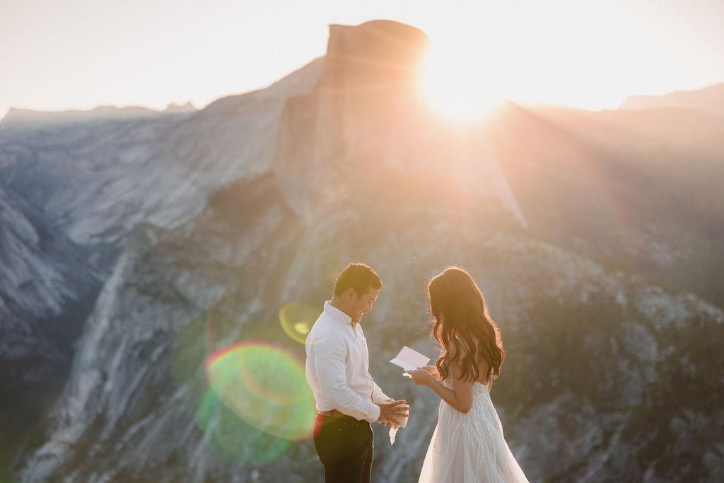 A couple stands on a rocky ledge during sunset; the man, wearing a white shirt and dark pants, holds the hand of the woman, who is in a flowing white dress. A mountain peak is visible in the background for their yosemite elopement photos