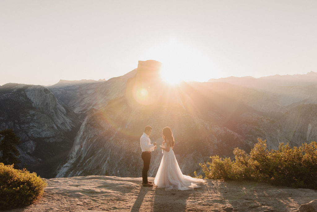 A couple stands on a rocky ledge during sunset; the man, wearing a white shirt and dark pants, holds the hand of the woman, who is in a flowing white dress. A mountain peak is visible in the background for their yosemite elopement photos