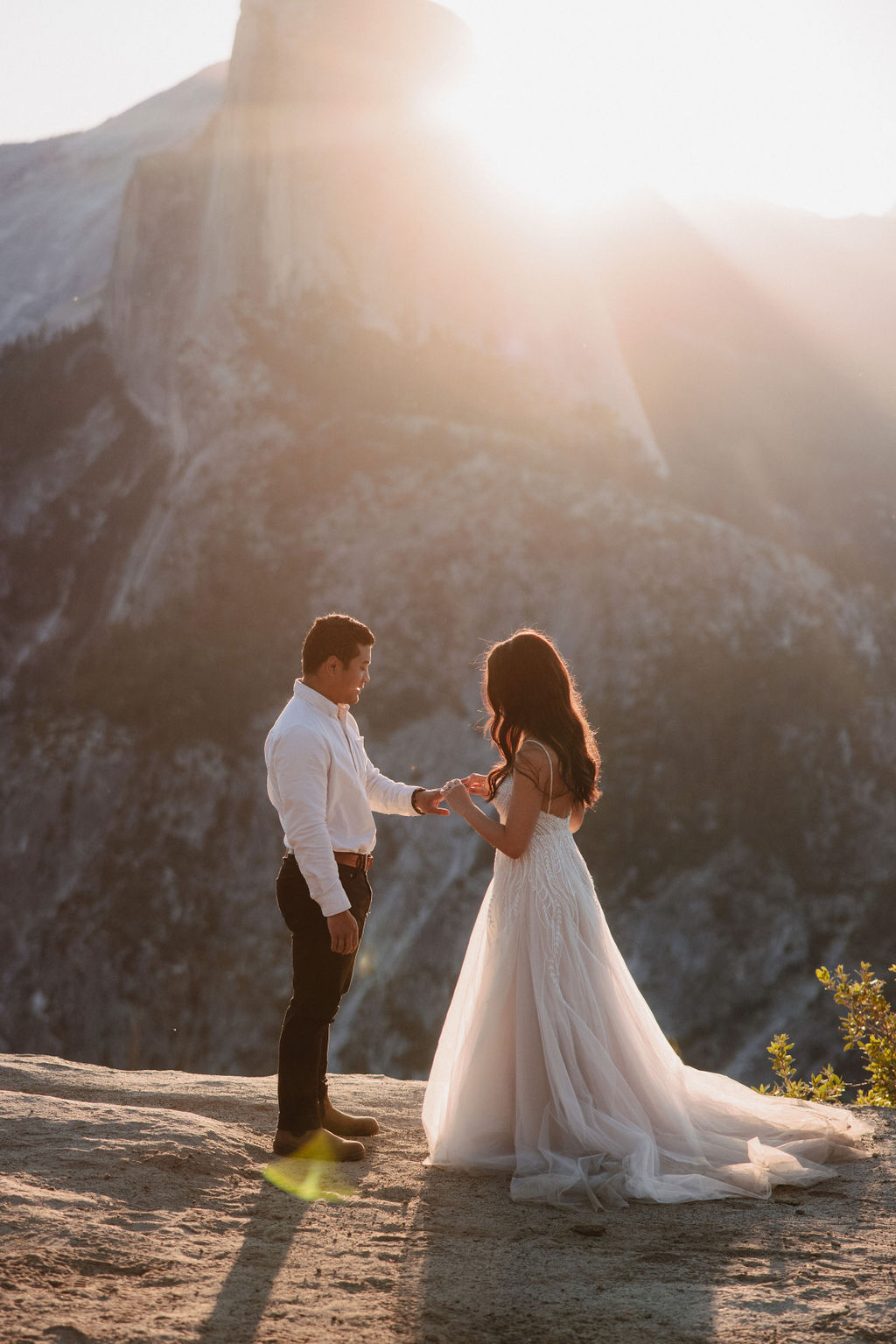 A couple stands on a rocky ledge during sunset; the man, wearing a white shirt and dark pants, holds the hand of the woman, who is in a flowing white dress. A mountain peak is visible in the background for their yosemite elopement photos