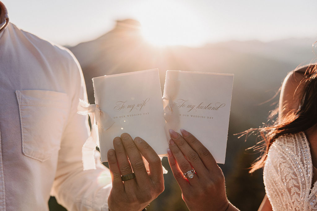 A couple stands on a rocky ledge during sunset; the man, wearing a white shirt and dark pants, holds the hand of the woman, who is in a flowing white dress. A mountain peak is visible in the background for their yosemite elopement photos