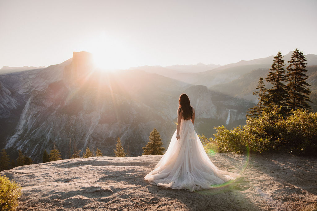 A bride in a flowing white dress stands on a rocky ledge at sunset, with a mountainous landscape and trees in the background for yosemite elopement photos