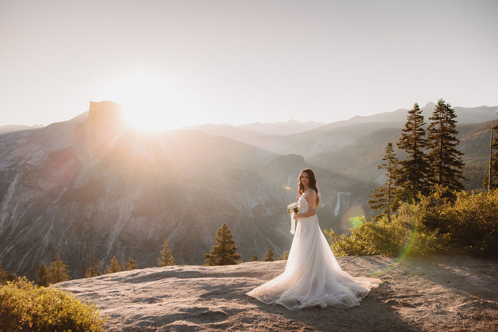 A bride in a flowing white dress stands on a rocky ledge at sunset, with a mountainous landscape and trees in the background for yosemite elopement photos