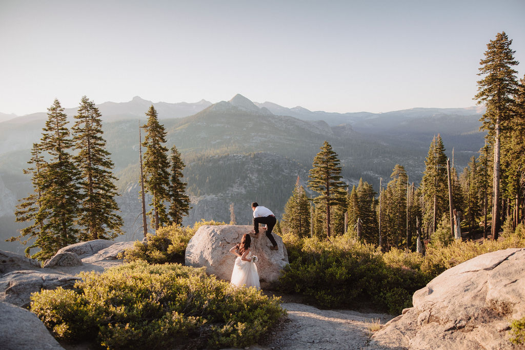 A couple stands on a rocky ledge during sunset; the man, wearing a white shirt and dark pants, holds the hand of the woman, who is in a flowing white dress. A mountain peak is visible in the background for their yosemite elopement photos