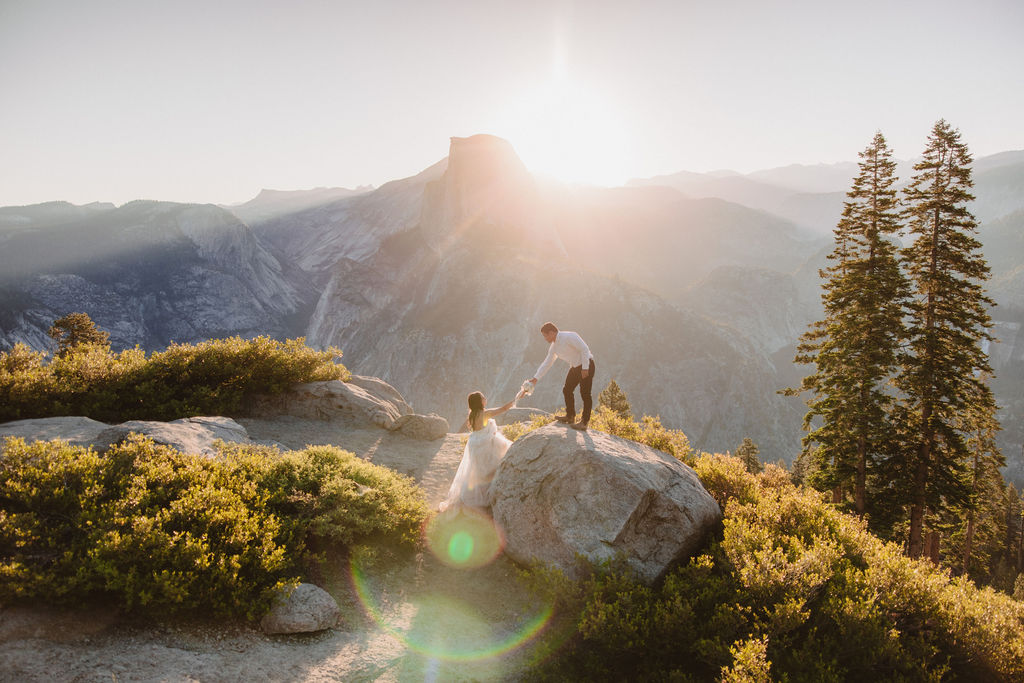 A couple stands on a rocky ledge during sunset; the man, wearing a white shirt and dark pants, holds the hand of the woman, who is in a flowing white dress. A mountain peak is visible in the background for their yosemite elopement photos