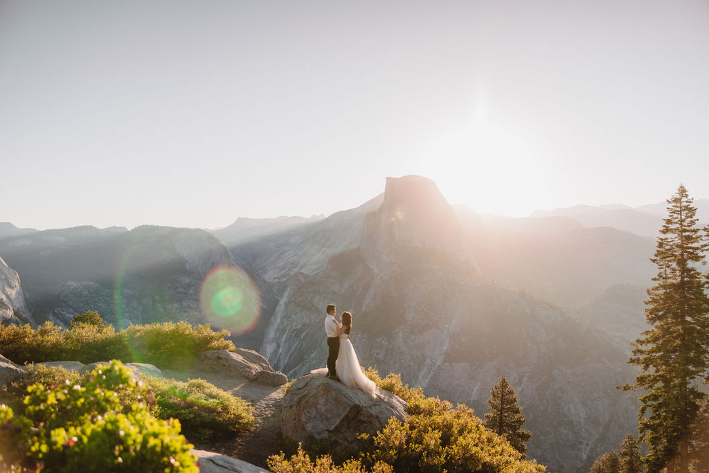 A couple stands on a rocky ledge during sunset; the man, wearing a white shirt and dark pants, holds the hand of the woman, who is in a flowing white dress. A mountain peak is visible in the background for their yosemite elopement photos