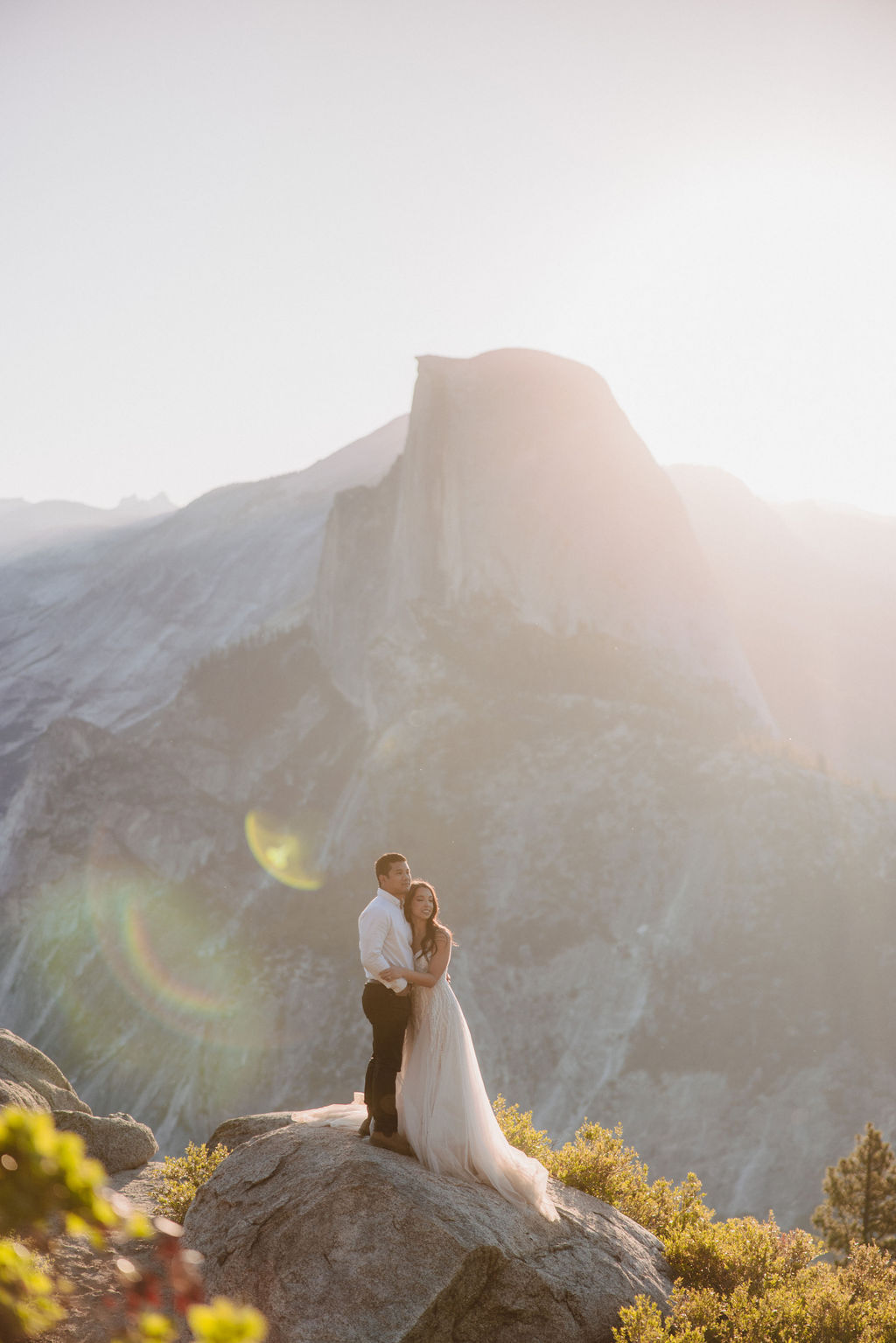 A couple stands on a rocky ledge during sunset; the man, wearing a white shirt and dark pants, holds the hand of the woman, who is in a flowing white dress. A mountain peak is visible in the background for their yosemite elopement photos