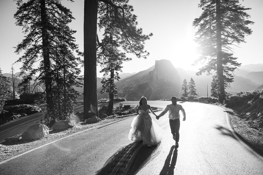 A bride and groom holding hands are walking down the middle of a road in a forested area with a pedestrian crossing sign visible.