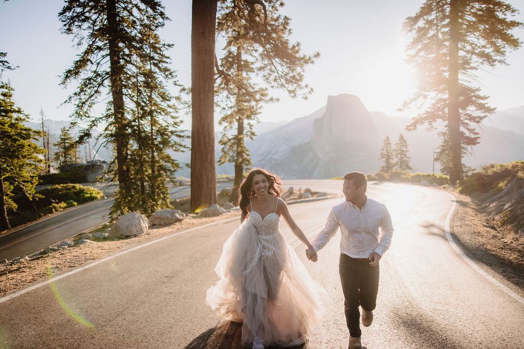 A bride and groom holding hands are walking down the middle of a road in a forested area with a pedestrian crossing sign visible.