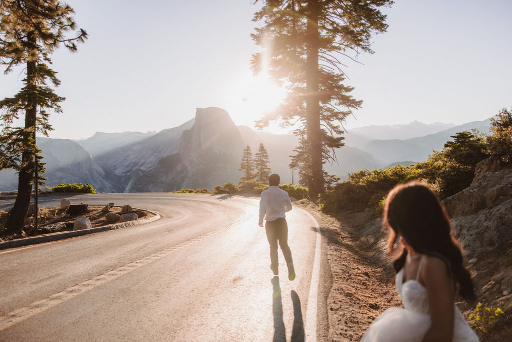 A bride and groom holding hands are walking down the middle of a road in a forested area with a pedestrian crossing sign visible.