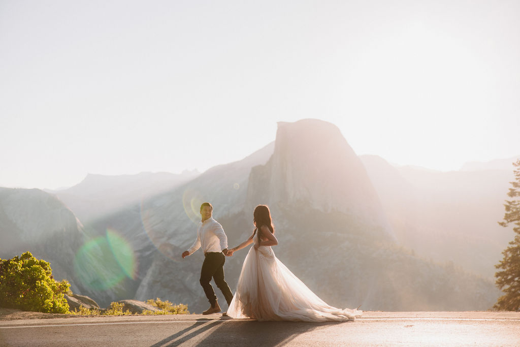 A couple stands on a rocky ledge during sunset; the man, wearing a white shirt and dark pants, holds the hand of the woman, who is in a flowing white dress. A mountain peak is visible in the background for their yosemite elopement photos