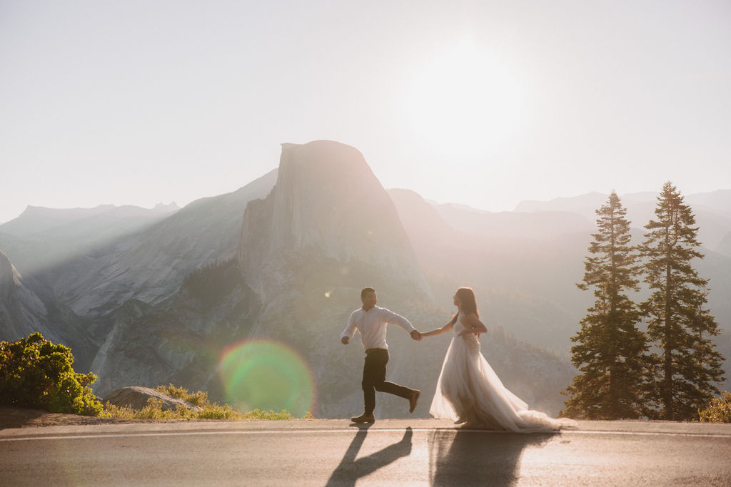 A couple stands on a rocky ledge during sunset; the man, wearing a white shirt and dark pants, holds the hand of the woman, who is in a flowing white dress. A mountain peak is visible in the background for their yosemite elopement photos