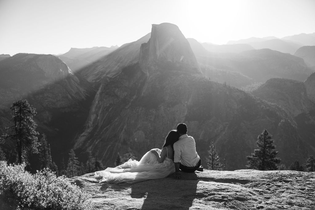 A couple stands on a rocky ledge during sunset; the man, wearing a white shirt and dark pants, holds the hand of the woman, who is in a flowing white dress. A mountain peak is visible in the background for their yosemite elopement photos