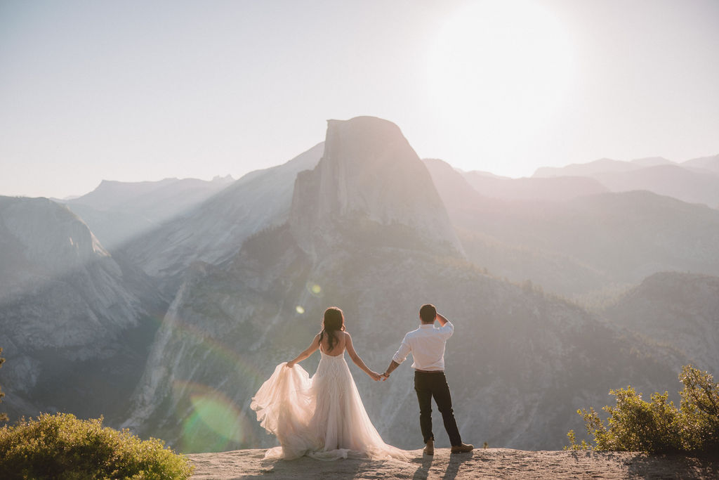 A couple stands on a rocky ledge during sunset; the man, wearing a white shirt and dark pants, holds the hand of the woman, who is in a flowing white dress. A mountain peak is visible in the background for their yosemite elopement photos