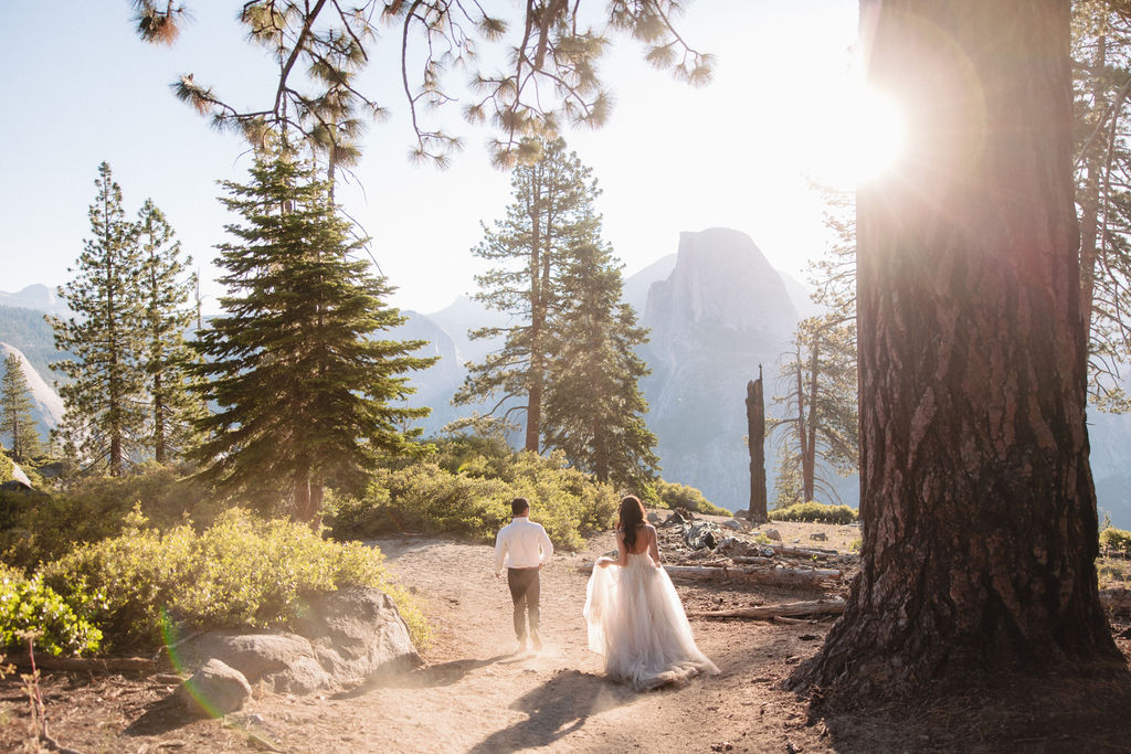 A couple walks along a forest path, with the woman in a white dress and the man in a white shirt and dark pants, surrounded by tall trees and sunlight filtering through.