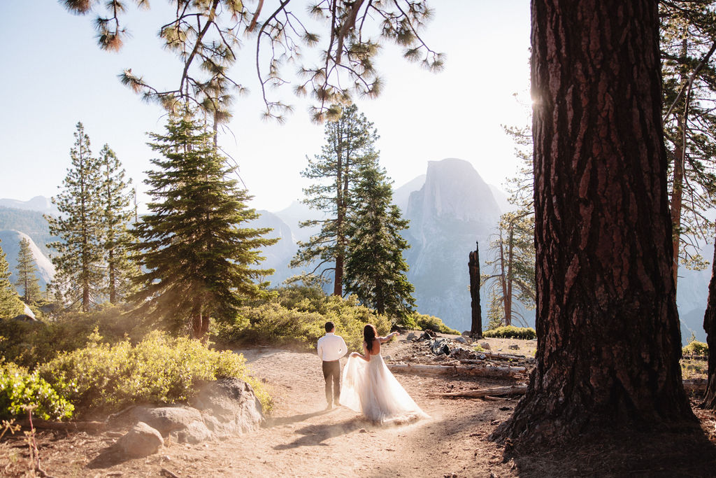 A couple walks along a forest path, with the woman in a white dress and the man in a white shirt and dark pants, surrounded by tall trees and sunlight filtering through.