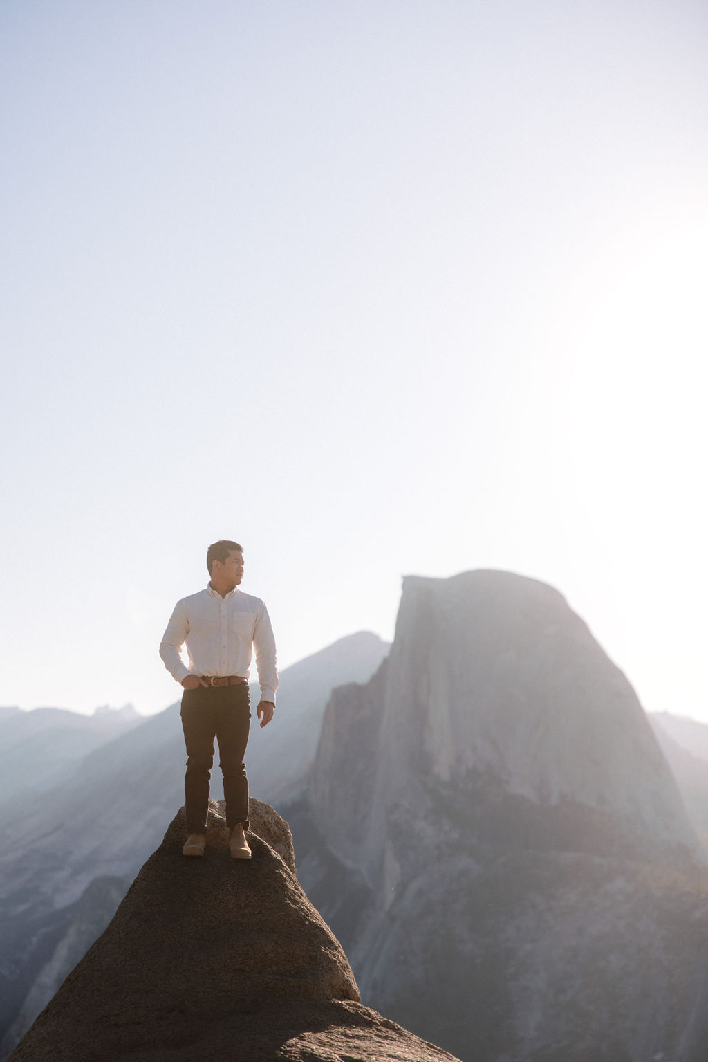 A couple stands on a rocky ledge during sunset; the man, wearing a white shirt and dark pants, holds the hand of the woman, who is in a flowing white dress. A mountain peak is visible in the background for their yosemite elopement photos