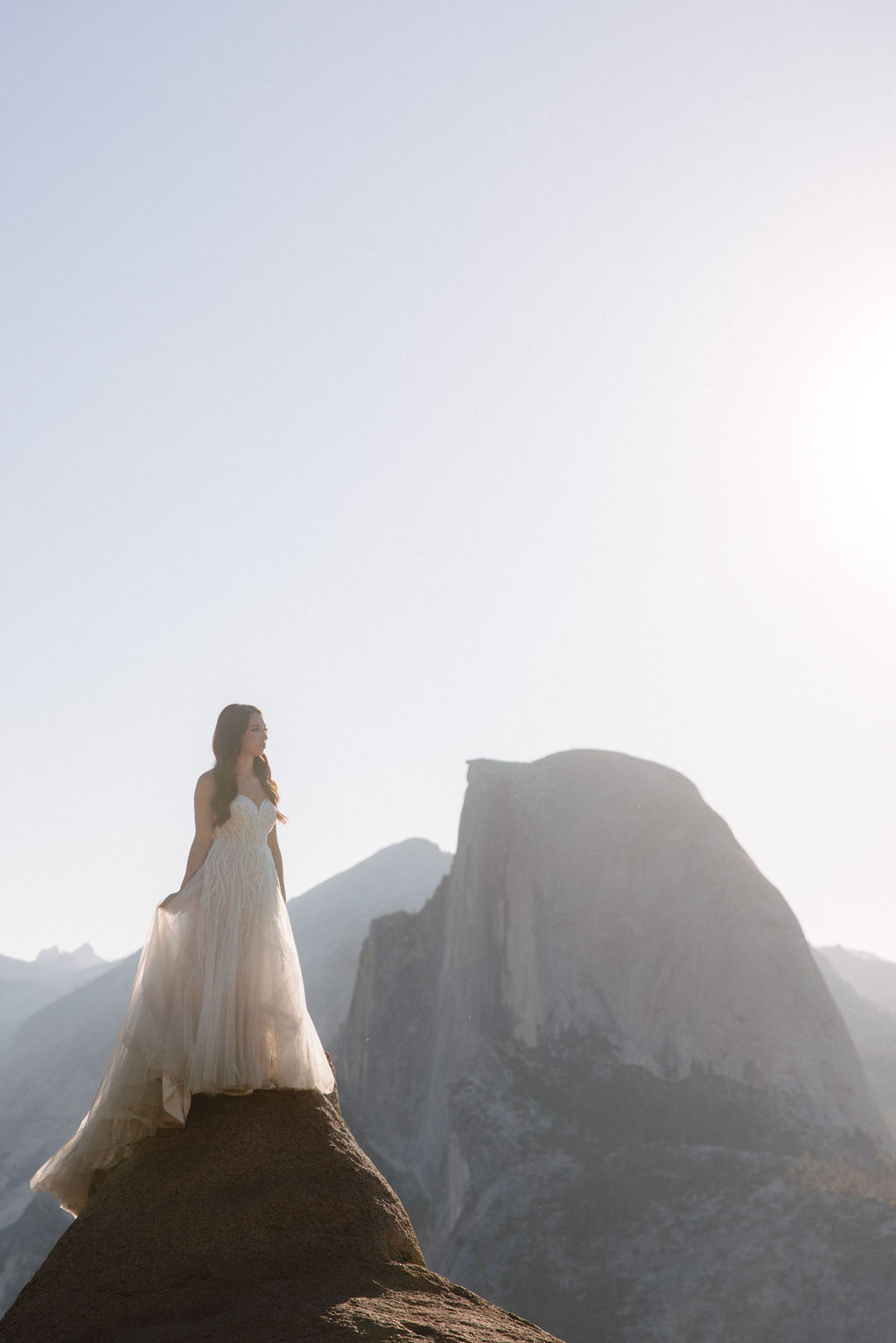 A couple stands on a rocky ledge during sunset; the man, wearing a white shirt and dark pants, holds the hand of the woman, who is in a flowing white dress. A mountain peak is visible in the background for their yosemite elopement photos