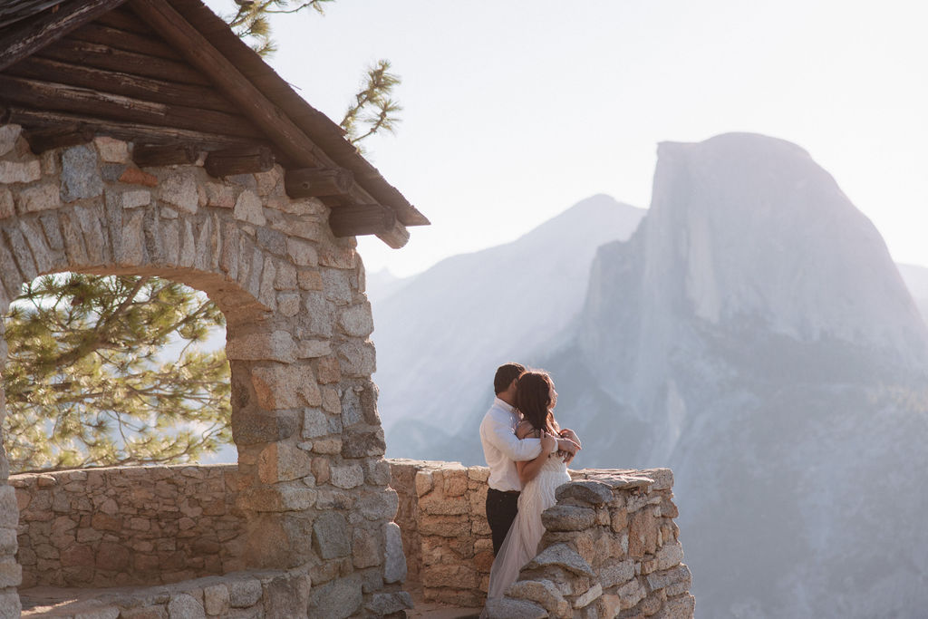 A couple embraces on a stone terrace with a wooden roof, with mountainous scenery in the background.