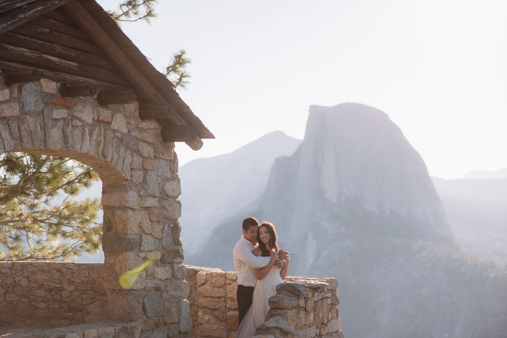 A couple embraces on a stone terrace with a wooden roof, with mountainous scenery in the background.