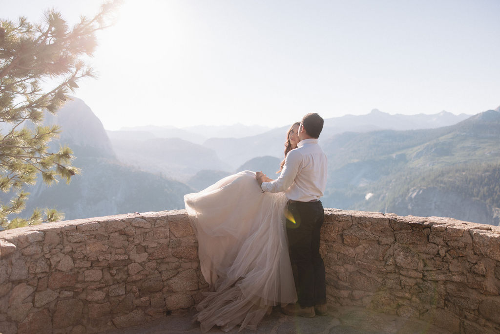 A couple embraces on a stone terrace with a wooden roof, with mountainous scenery in the background.