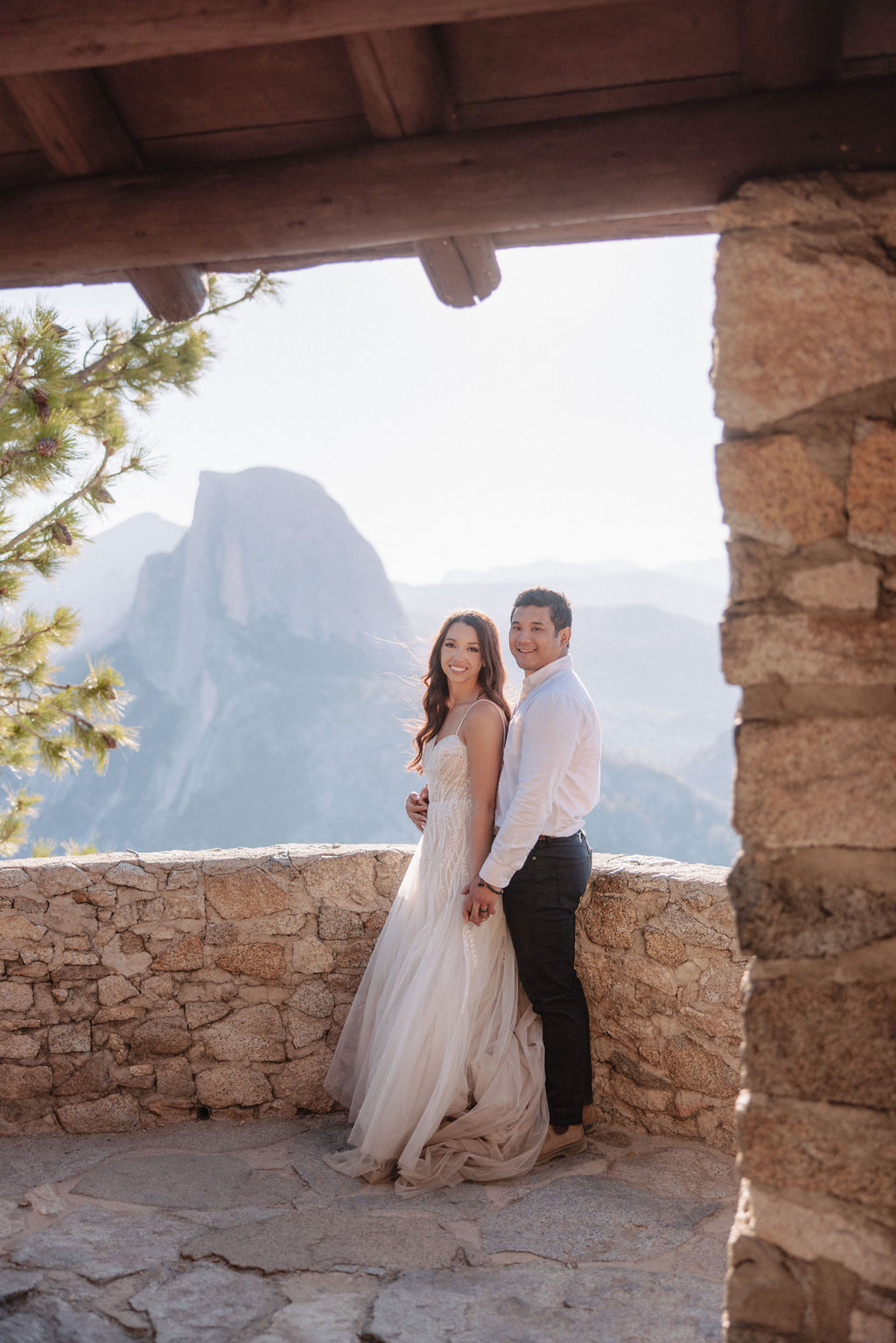 A couple embraces on a stone terrace with a wooden roof, with mountainous scenery in the background.