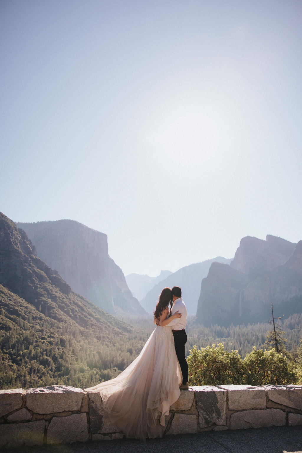 A couple embraces on a stone terrace with a wooden roof, with mountainous scenery in the background.