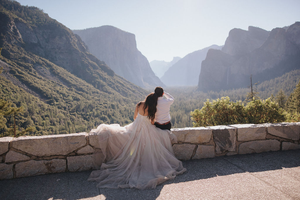 A couple embraces on a stone terrace with a wooden roof, with mountainous scenery in the background.