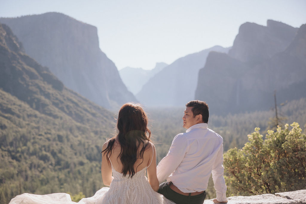 A couple embraces on a stone terrace with a wooden roof, with mountainous scenery in the background.