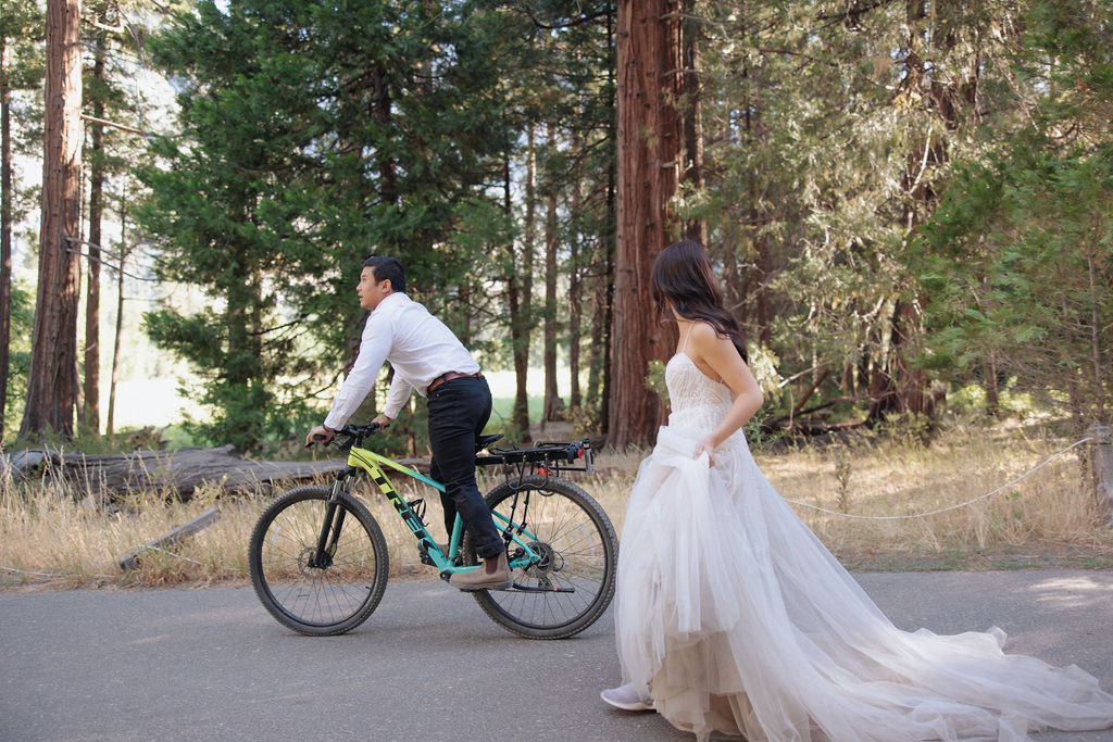 A bride in a white dress stands on a forest trail, looking at a man riding a bicycle ahead of her for their Yosemite elopement photos