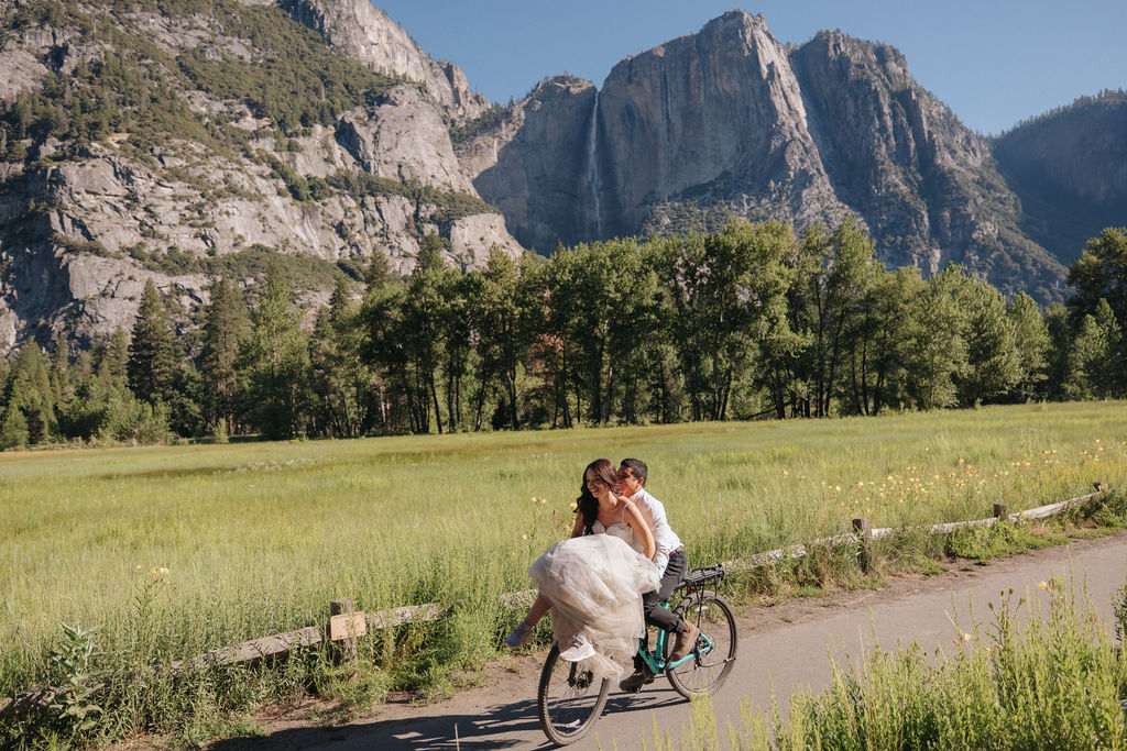 A bride in a white dress stands on a forest trail, looking at a man riding a bicycle ahead of her for their Yosemite elopement photos