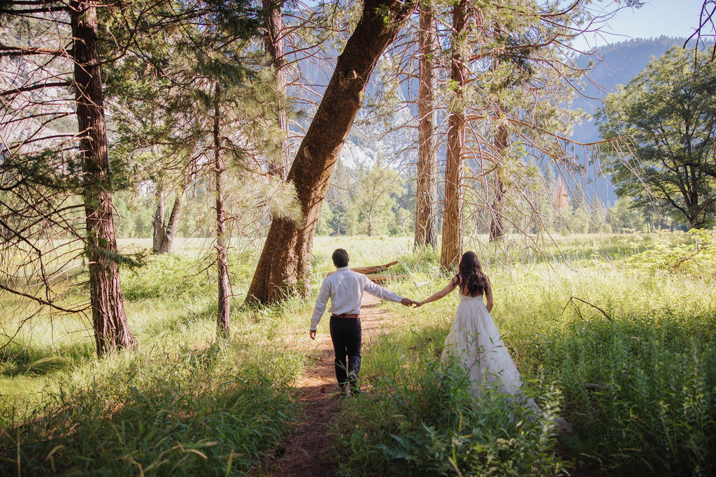 A couple stands in a meadow with a mountainous landscape in the background. The bride is twirling in her dress while the groom looks on at Yosemite elopement photos