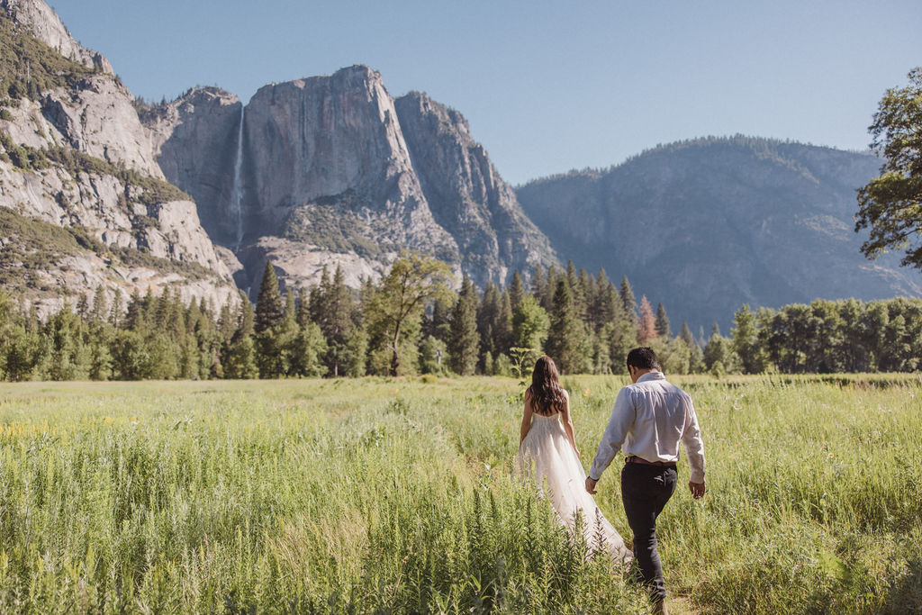 A couple stands in a meadow with a mountainous landscape in the background. The bride is twirling in her dress while the groom looks on at Yosemite elopement photos