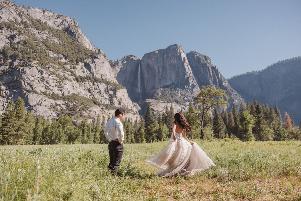 A couple stands in a meadow with a mountainous landscape in the background. The bride is twirling in her dress while the groom looks on at Yosemite elopement photos