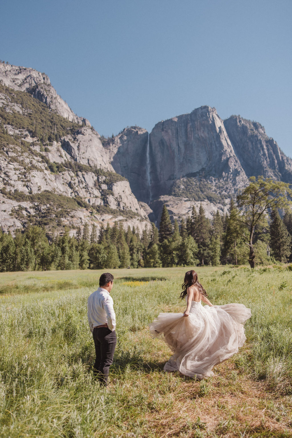 A couple stands in a meadow with a mountainous landscape in the background. The bride is twirling in her dress while the groom looks on at Yosemite elopement photos