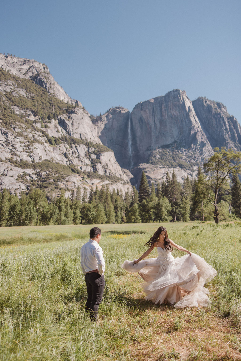 A couple stands in a meadow with a mountainous landscape in the background. The bride is twirling in her dress while the groom looks on at Yosemite elopement photos