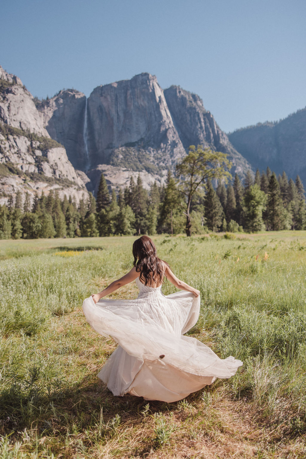 A couple stands in a meadow with a mountainous landscape in the background. The bride is twirling in her dress while the groom looks on at Yosemite elopement photos