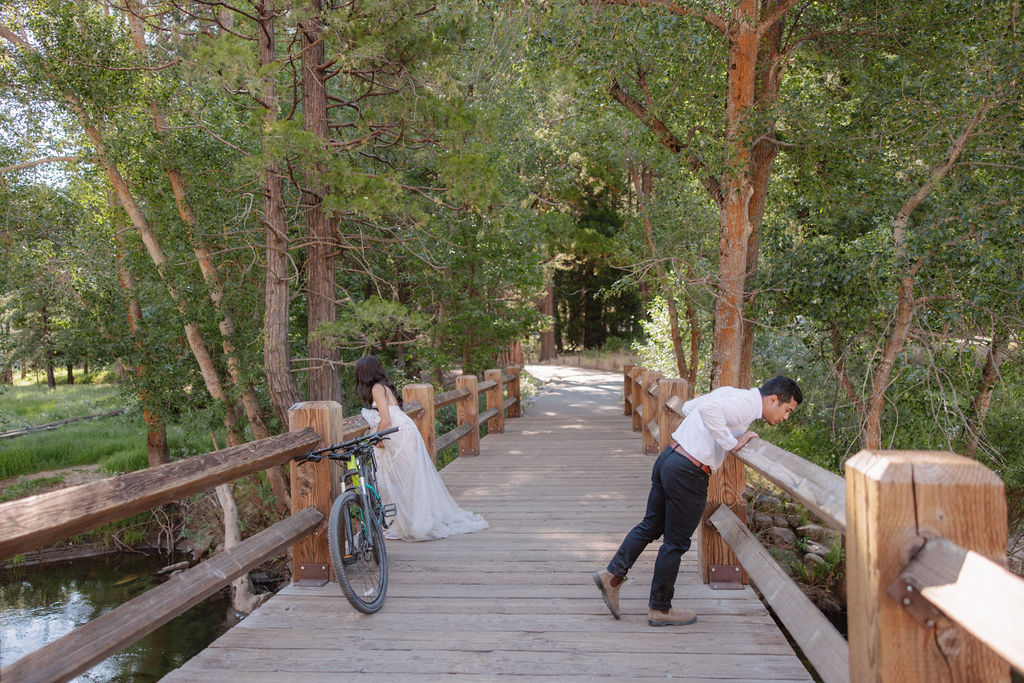 A bride in a white dress and a groom in a white shirt, both standing on a wooden bridge in a forested area; a bicycle is leaned against the railing.