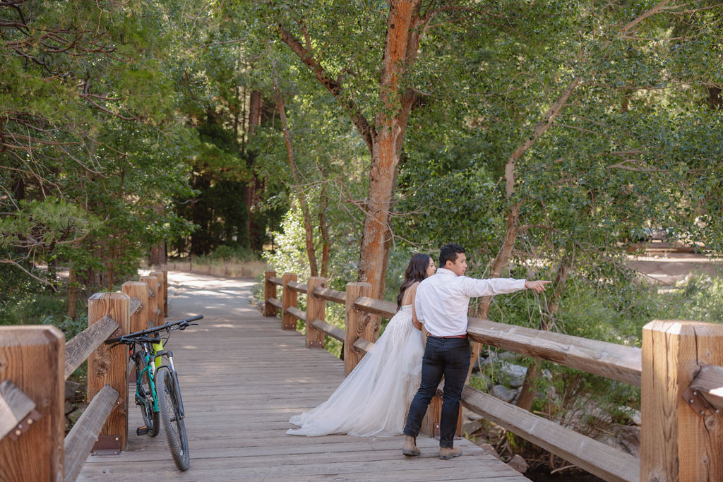 A bride in a white dress and a groom in a white shirt, both standing on a wooden bridge in a forested area; a bicycle is leaned against the railing.