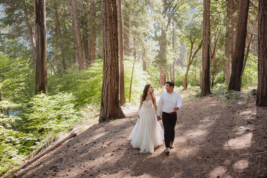 Bride in a white dress and groom in a white shirt standing on a large fallen tree with a forest and rocky mountain in the background.