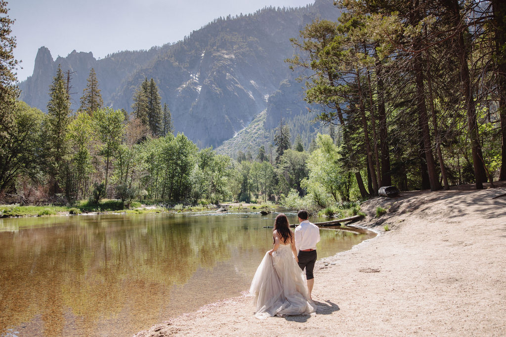 A woman in a white dress and a man in dress clothes wade in a shallow, clear river surrounded by trees and greenery.