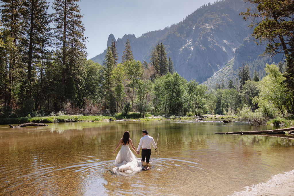 A woman in a white dress and a man in dress clothes wade in a shallow, clear river surrounded by trees and greenery.