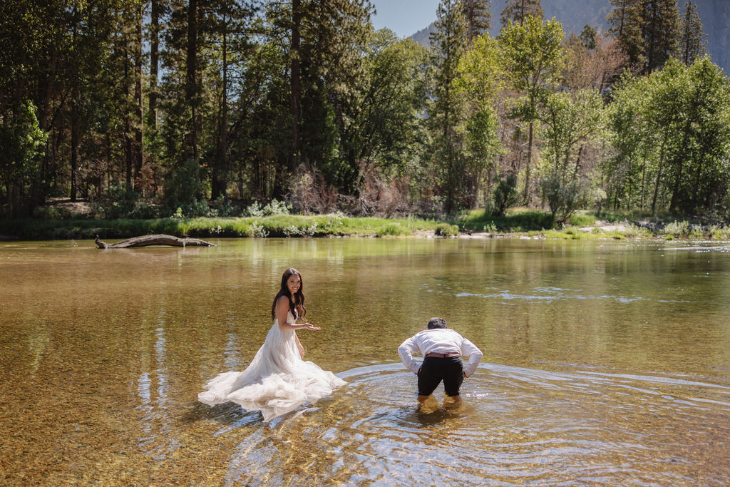 A woman in a white dress and a man in dress clothes wade in a shallow, clear river surrounded by trees and greenery.