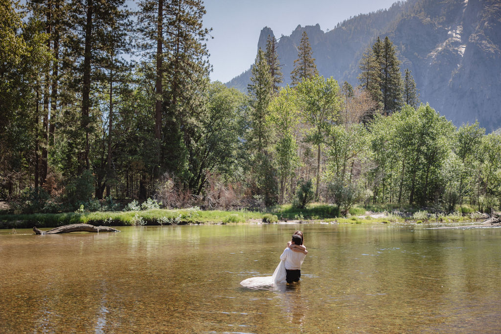 A woman in a white dress and a man in dress clothes wade in a shallow, clear river surrounded by trees and greenery.